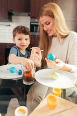 Obraz na płótnie Canvas Young beautiful blonde mother and her cute four year old son having fun while painting eggs for Easter at home kitchen, smiling, developing creative abilities.
