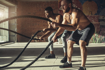 Muscular half-naked body athletes doing some crossfit exercises with a rope indoor, preparing to...