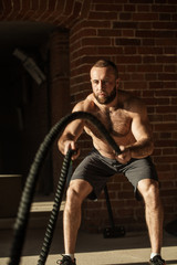 Fototapeta na wymiar Muscular powerful determined man training with rope in functional training in outdoor gym with brick walls, strengthen the muscles of the shoulders, which is important for boxers and swimmers