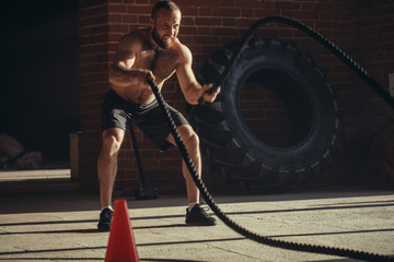 Fototapeta na wymiar Muscular powerful determined man training with rope in functional training in outdoor gym with brick walls, strengthen the muscles of the shoulders, which is important for boxers and swimmers
