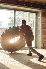 Obraz na płótnie Canvas Rear view of African american crossfit man pushing hard with dedication and determination heavy tire in gym, training strength and endurance