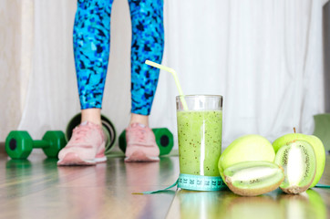 Concept of sport and healthy lifestyle.Female legs standing on wooden floor near the sport equipment-mat and dumbbells.Fresh green diet fruits,cocktail and measuring tape