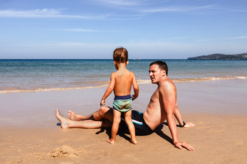 Unrecognizable little boy in swimwear standing on sandy beach near adult man on sunny day on resort