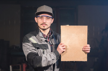 Portrait of a young carpenter in overalls and goggles with a mock up board holding a blank sign in his hands in his home workshop