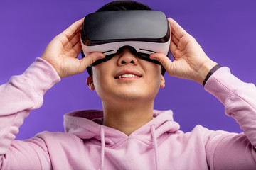 Asian young man dressed in white hoodie using virtual reality headset isolated over violet background