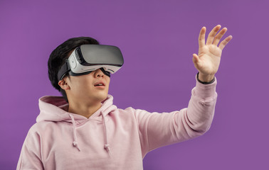 Asian young man dressed in white hoodie using virtual reality headset isolated over violet background