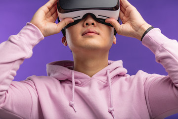 Asian young man dressed in white hoodie using virtual reality headset isolated over violet...