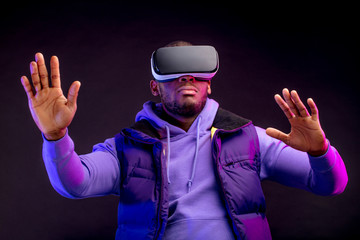 African professional boxer in casual purple wear looking through the competition with his participation, using VR glasses, studying mistakes and tactic of opponent. Dark background.