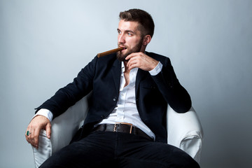 Portrait of a bearded businessman with a cigar, who is sitting in a white armchair and relaxing