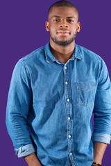 Studio portrait of confident charismatic young african-blogger dressed in casual jeans shirt holding hands in pockets looking at camera over purple background.