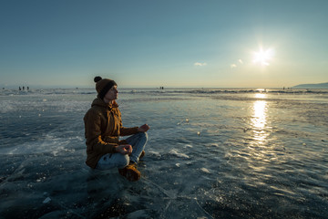 Fototapeta premium Tourist with a backpack meditates sitting in Lotus position on the ice of lake Baikal. Relaxation and freedom of nature
