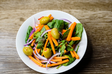 Freshly made vegetable salad with carrot, olives, spinach and onion in a bowl on wooden background.