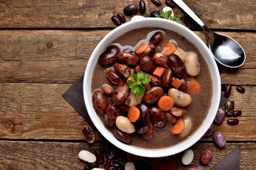 Close-up of full bowl of hot bean soup with large beans on cutting board, carrots, parsley, marjoram, spoon in background - top view