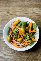 Freshly made vegetable salad with carrot, olives, spinach and onion in a bowl on wooden background.
