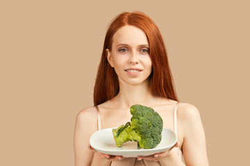 Young skinny woman with ginger hair and starved face in underwear looking at camera holding plate with raw broccoli, happy to eat something