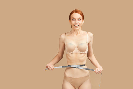 Woman suffering eating disorders anorexia or bulimia in nude bra and panties celebrates diet results, measuring waist with tape over beige background