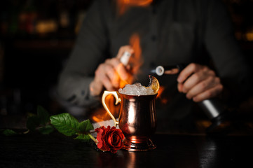 Professional bartender spraying on the delicious cocktail in the golden cup and flaming it with a burner