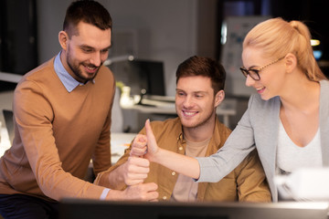 business, success and teamwork concept - happy coworkers with computer working late at night office making thumbs up gesture