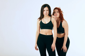 Two caucasian lesbian women posing together in black sportswear on white, red-haired girl leaning with a shoulder to her brunette girlfriend