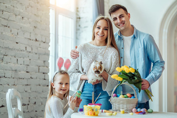 Happy easter!Dad holds a bouquet of tulips, mom holds a home decorative rabbit, daughter paints eggs and looking at camera.Celebration concept.