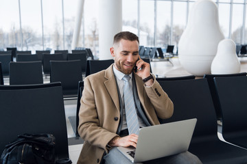 Happy businessman working on the laptop and talking on cellphone at the airport waiting lounge. Handsome caucasian businessman at waiting room in airport terminal