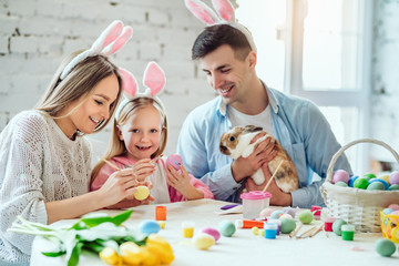 We love beautiful traditions.Mom and daughter paint eggs, dad holds a home decorative rabbit.
