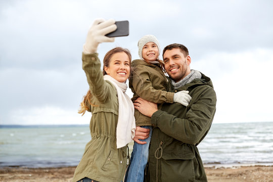 family, leisure and people concept - happy mother, father and little daughter taking selfie by smartphone on autumn beach