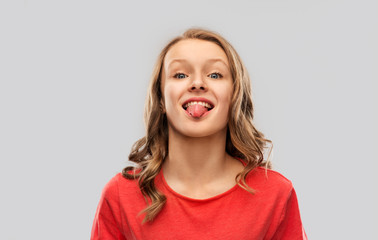 gesture, expression and people concept - funny teenage girl with long hair in red t-shirt showing tongue over grey background