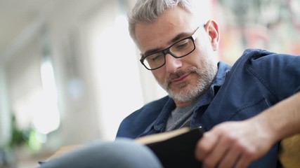 Mature man wearing glasses reading book at home - Powered by Adobe