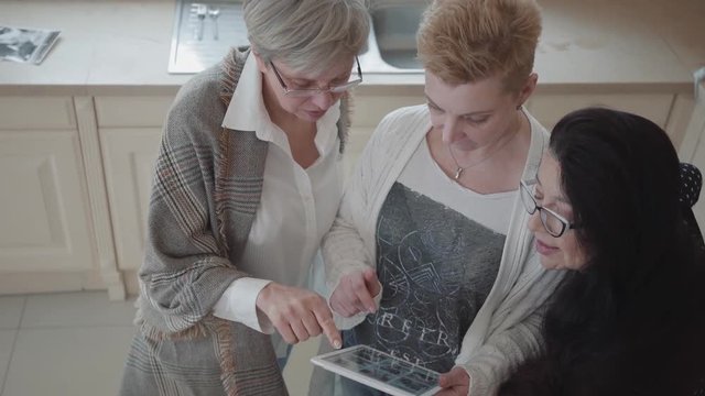 Senior woman showing to her friends photos on tablet. Group of three middle aged mature women communicating chatting spending time in the kitchen at home with wine and fruits. Top view