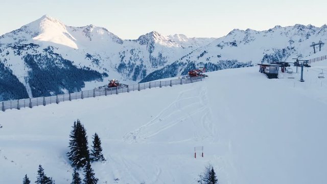 Two snowcats are going up a mountain just before dark. The drone flies towards them and the snow covered mountains in the background.