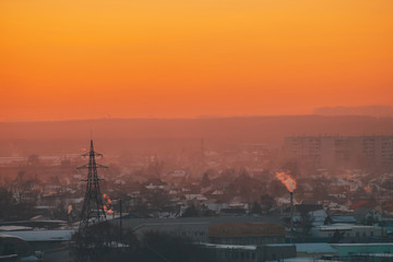 Naklejka premium Power lines in city on dawn. Silhouettes of urban buildings among smog on sunrise. Cables of high voltage on warm orange yellow sky. Power industry at sunset. City power supply. Mist urban background.