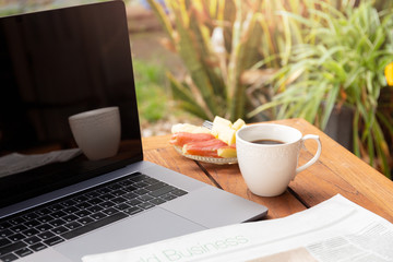 Coffee mug and news paper with fresh fruit and laptop on wooden table.