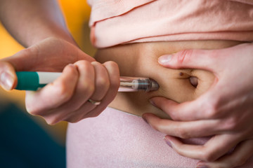 Woman making hormonal therapy injection into her belly. Close up syringe pen. Person in bedroom, in...