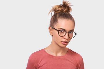 Attractive blonde young woman wears spectacles and casual pink blouse, looks seriously directly into camera, poses against white studio background. People emotions concept