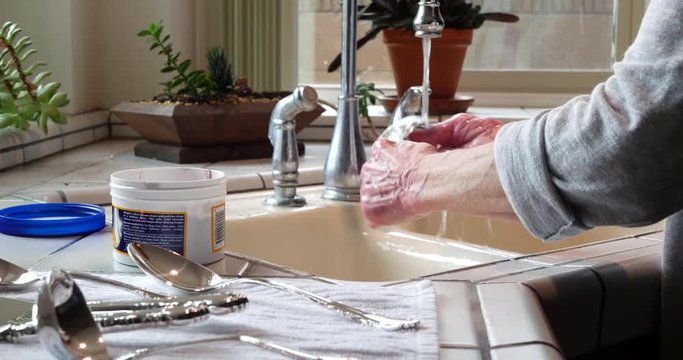 The hands of an elderly woman cleaning and polishing a piece of silverware and rinsing it in the kitchen sink to remove tarnish.