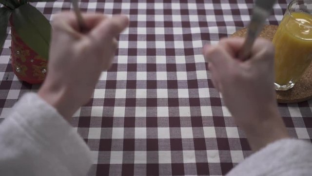Man's hand knocking on the table with his fists, demanding food, holding fork and knife close up. Orange juice glass and vase are on the table with checkered tablecloth. First person shooting