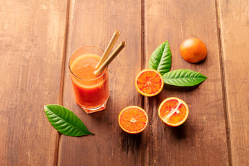 A glass of fresh orange juice with blood oranges, green leaves, and two bamboo straws, on a dark...