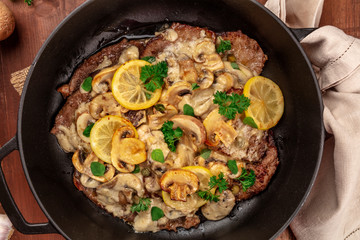 A closeup photo of scallopine di vitello, veal scallopini, a traditional Italian dish with a mushroom sauce, lemons, and parsley, shot from above in a braiser