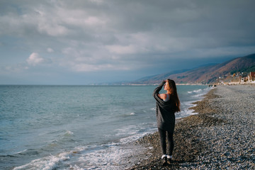 Girl in grey cardigan walking alone on empty pebble seashore in cloudy cold weather. Lonely woman freedom concept