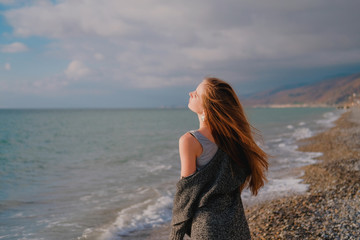 Obraz premium Girl in grey cardigan walking alone on empty pebble seashore in cloudy cold weather. Lonely woman freedom concept