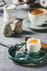Breakfast with cup of coffee and soft boiled egg, served in green ceramic egg cup with salt, pepper and toasted bread, jug of cream and cotton flowers over grey blue table.