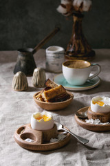 Breakfast with cup of coffee and soft boiled egg, served in wooden egg cup with salt, pepper and toasted bread, jug of cream and cotton flowers over linen tablecloth.
