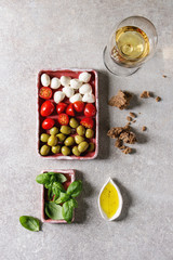 Mozzarella, cherry tomatoes, olives antipasto appetizers served in pink ceramic rectangular plates with olive oil, glass of white wine, basil, rye bread over grey texture background. Flat lay, space