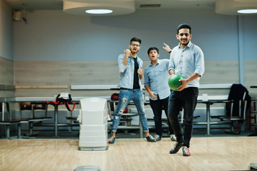 Fototapeta premium South asian man in jeans shirt standing at bowling alley with ball on hands. Guy is preparing for a throw. Friends support him loudly.