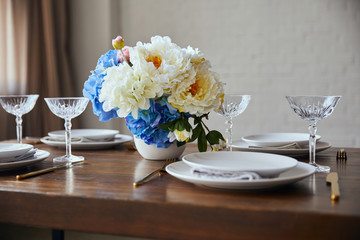 white plates, cutlery, crystal glasses and bouquet in vase on wooden table at home