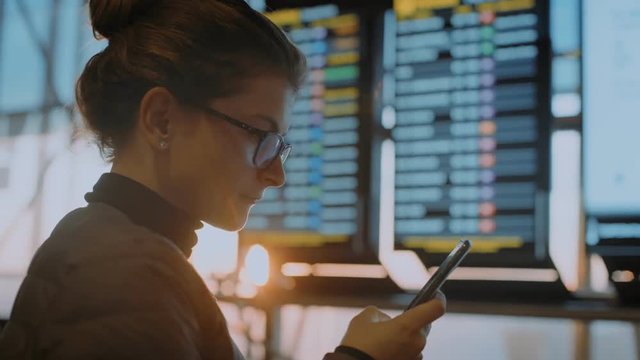 Young woman in casual business outfit during work or vacation trip stand in airport terminal or busy logistics international hub, looks for flight information with electronic ticket on smartphone