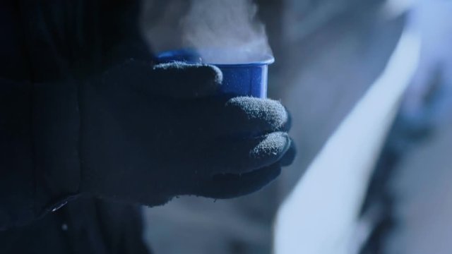 Slow motion close up of male or female hands in cotton gloves or mittens hold camping mug or cup with steaming hot beverage, tea or coffee on cold freezing winter day on outdoor adventure