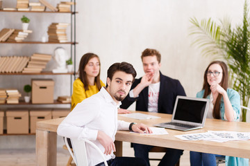 Fototapeta premium Portrait of handsome successful businessman in white shirt sitting at conference table against the background of blurry employees of his company