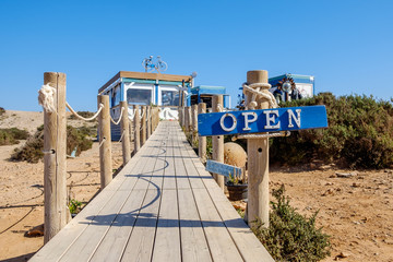 Colourful 'Open' sign, at the beginning of a boardwalk, leading to a rustic restaurant, on the mediterranean coast